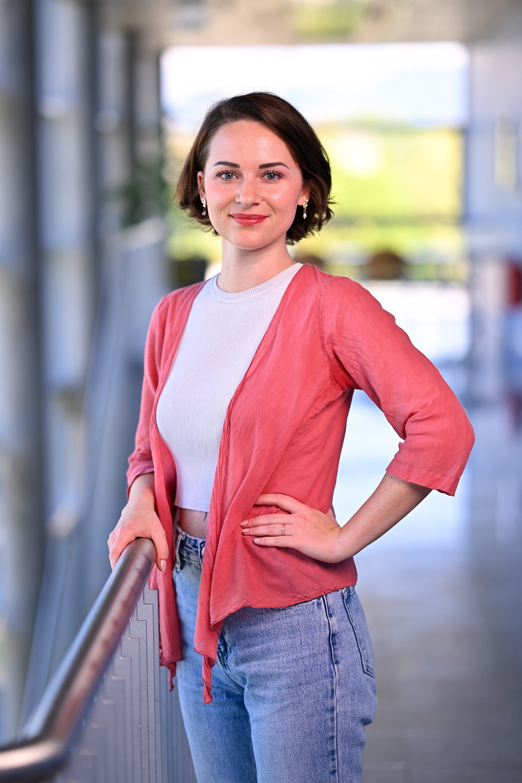 Antonia hat auf diesem Bild kinnlange braune, glatte Haare, eine rote dünne Jacke und darunter ein weißes Oberteil an. Sie steht in einem Gang auf der Fachhochschule Kärnten.